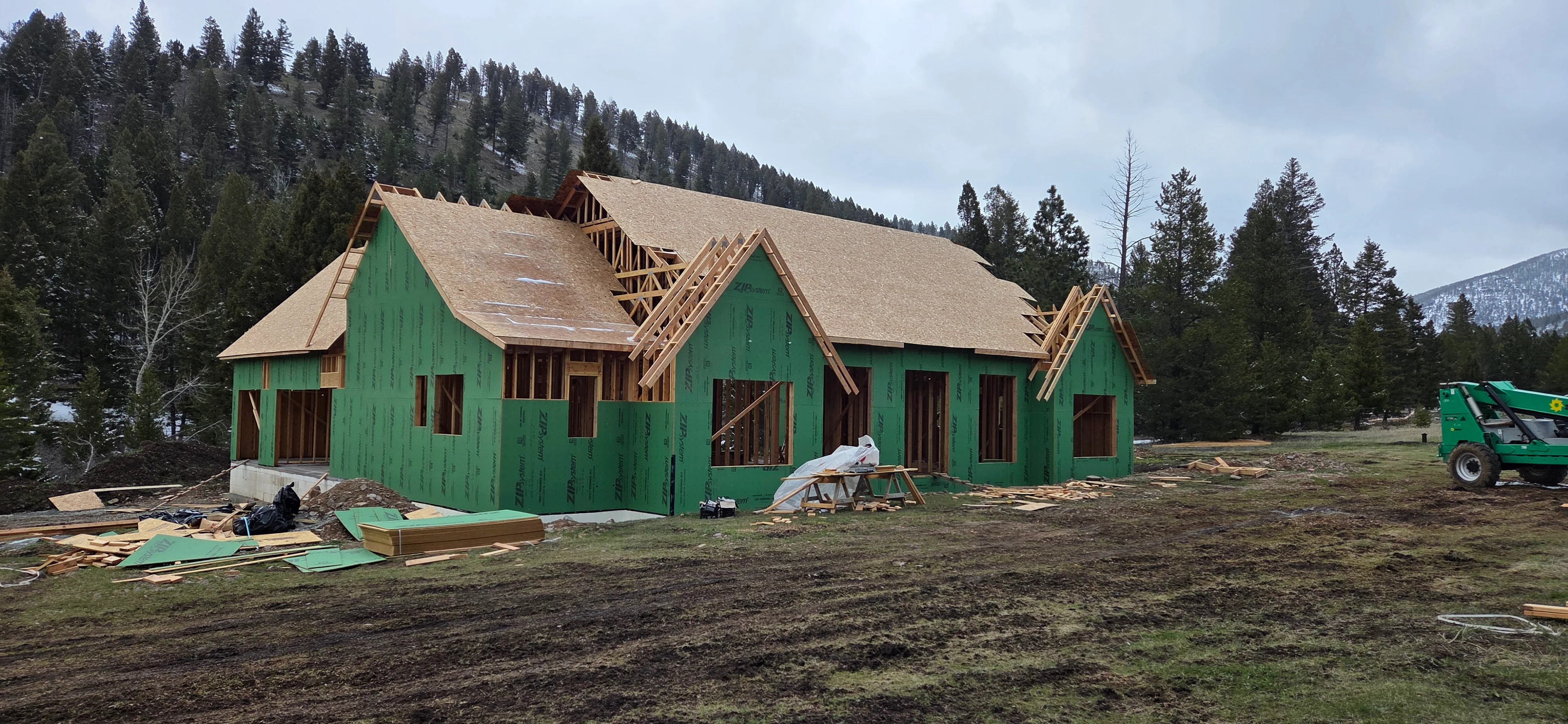 Home framing with green house wrap, exposed wood trusses, and composite shingle roofing in progress