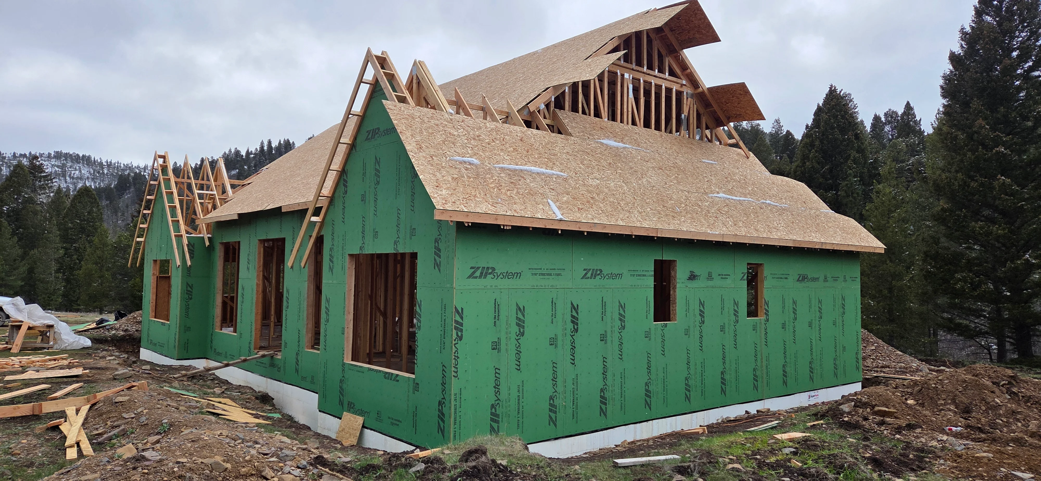 Wood-framed home with green weather wrap, complex gable roof framing, and asphalt shingle installation
