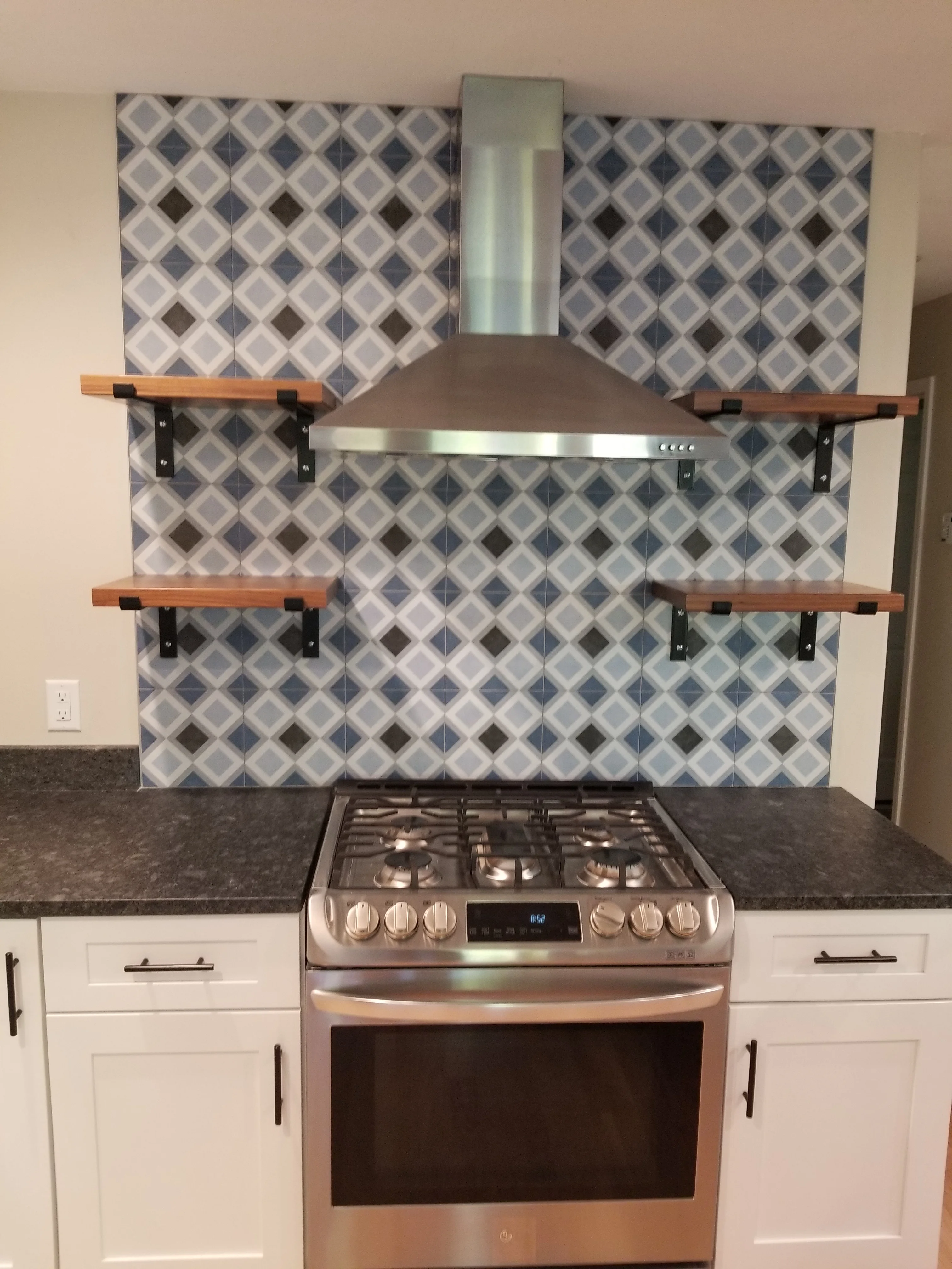 Geometric tile backsplash with stainless steel range hood and wooden open shelves above gas range