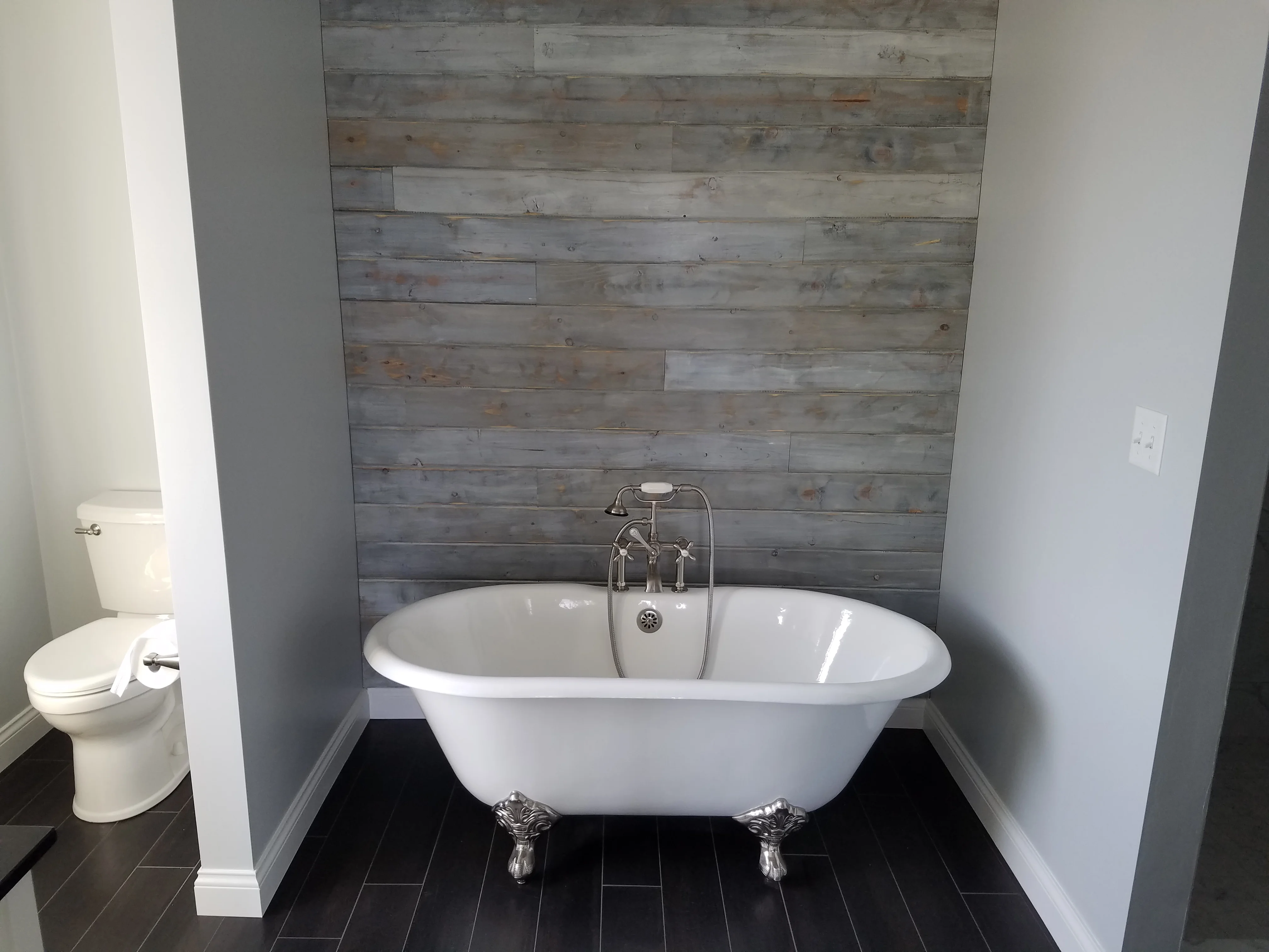 Freestanding clawfoot tub with gray shiplap accent wall, chrome fixtures, black tile floor